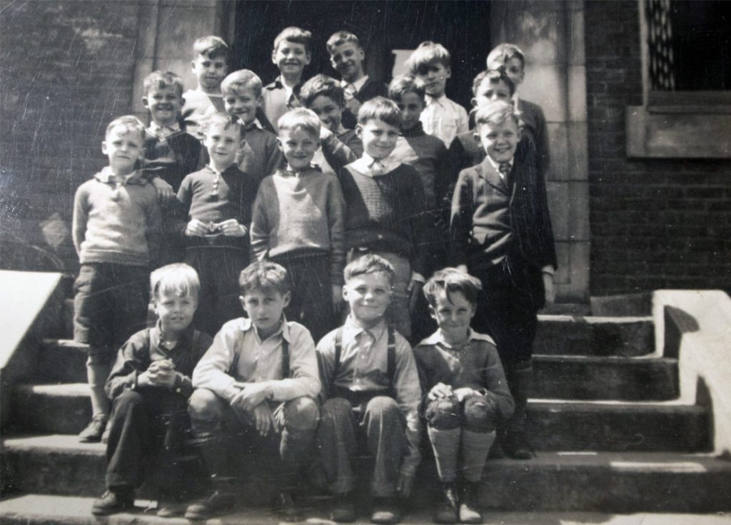 Nineteen young boys sit for a group photo on stairs outside a brick building.