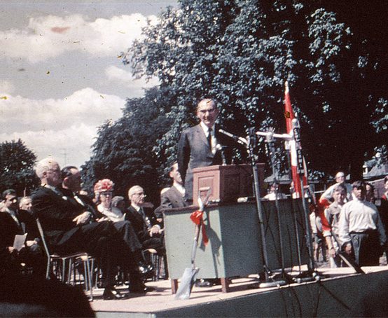 Man speaking at podium surrounded by microphones on a stage with other officials before an audience outside.