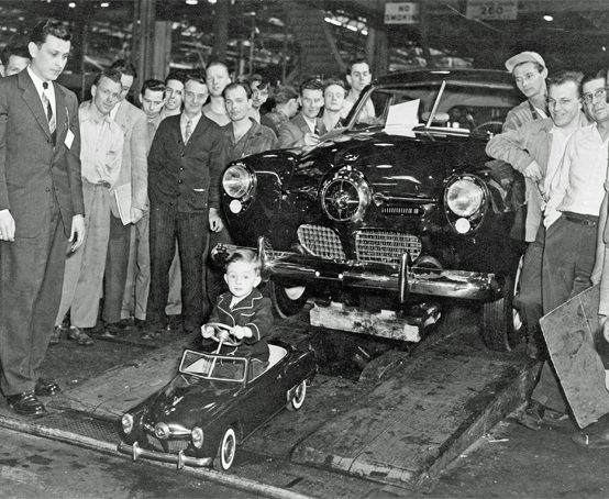Studebaker Bullet Nose Pedal Car driven by a little boy, leads a full size Studebaker off the assembly line.