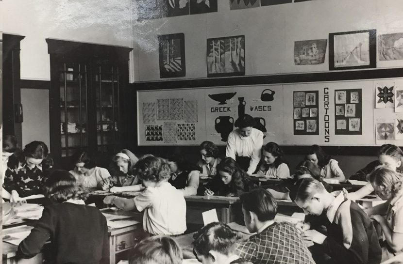 Male and female students work at their desks as the teacher assists.