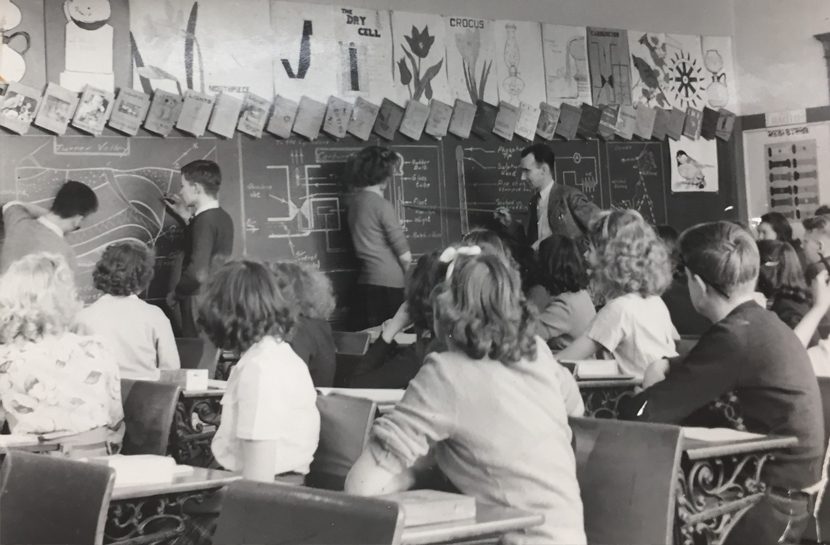 Male and female students look on as their peers work with the teacher at the blackboard.