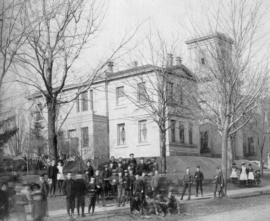People gather on the grounds of a large Georgian-style building with bell tower, and mature trees.