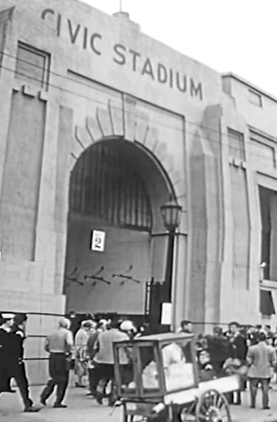 People walk around outside arched gate number two of the massive concrete facade of Civic Stadium.