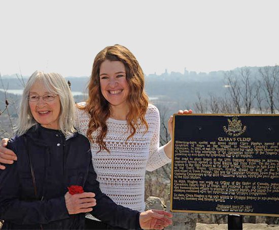 Two smiling woman standing near commemorative plaque.