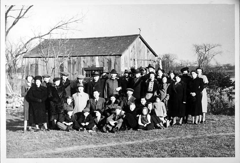 A large group of well dressed men, women and children pose in a rural area near a small wooden shed.