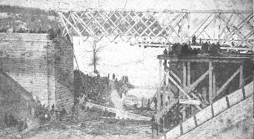 A train partially submerged in icy canal water after having broken through a trestle bridge draws a panicked crowd.