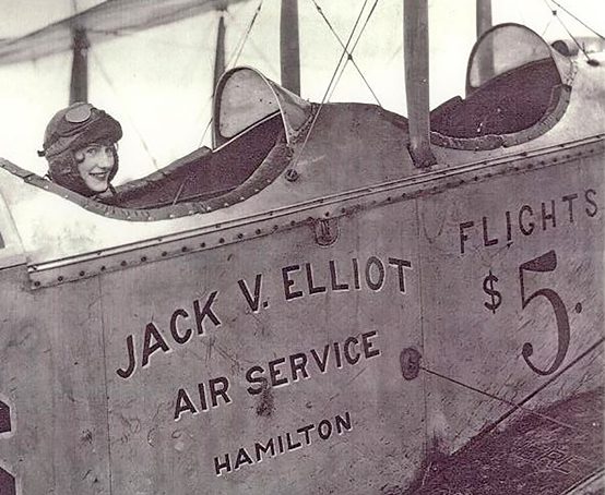 Woman wearing aviator hat and goggles sits in pilot seat of a plane marked Jack V. Elliot Air Service Hamilton.