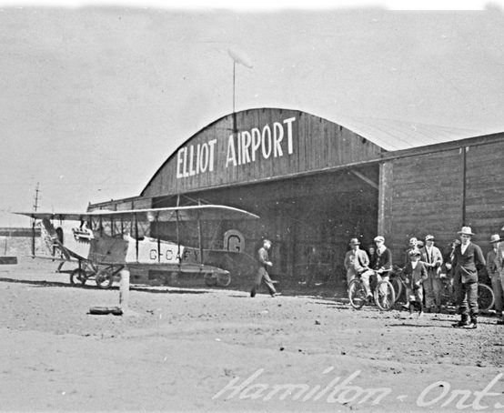 A biplane sits in the entrance of a hangar as people gather at the Elliot Airport Hamilton.