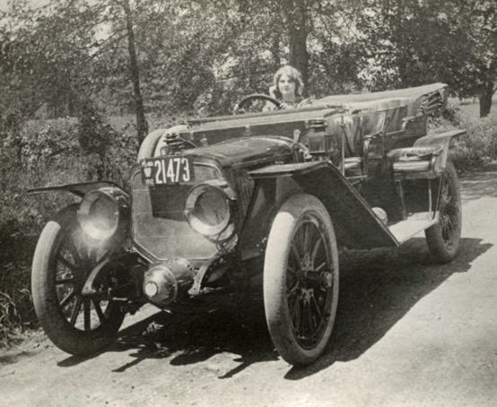 A vintage photo of woman sitting in a car parked at the side of a dirt road.