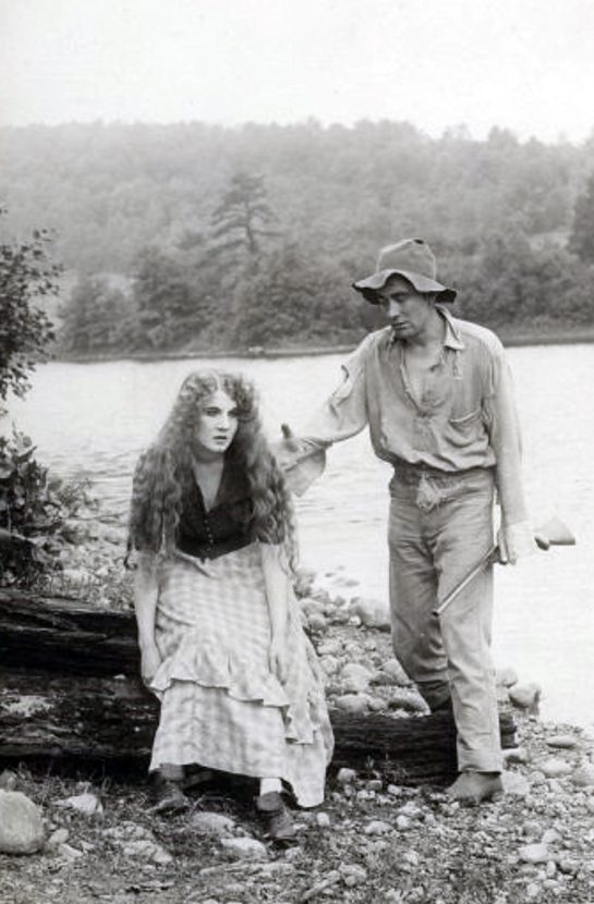 A man with a rifle reaches out to a distraught woman sitting on a log near a river bank.