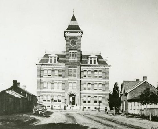 Large three story boxy stone building with bell tower and ornamental roof cresting. .