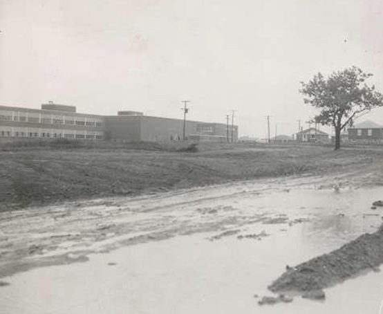 Street view of a modern brick building surrounded by unpaved muddy roads 1955.