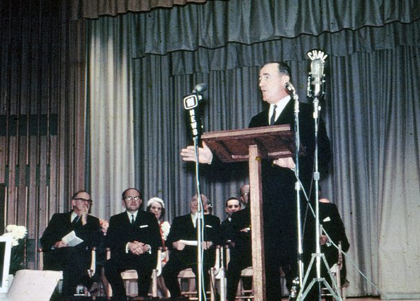 Man speaking while standing at podium surrounded by microphones on a stage with other officials.