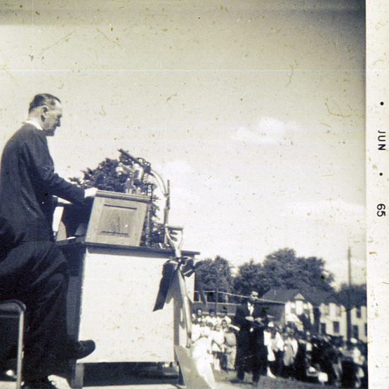 Man speaking at podium surrounded by microphones on a stage with other officials before an audience outside.