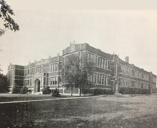 Street view of boxy two story building with many windows and a grand arched entrance.