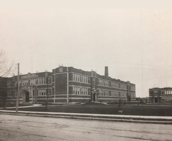 Street view of boxy two story building with many windows and a grand arched entrance.