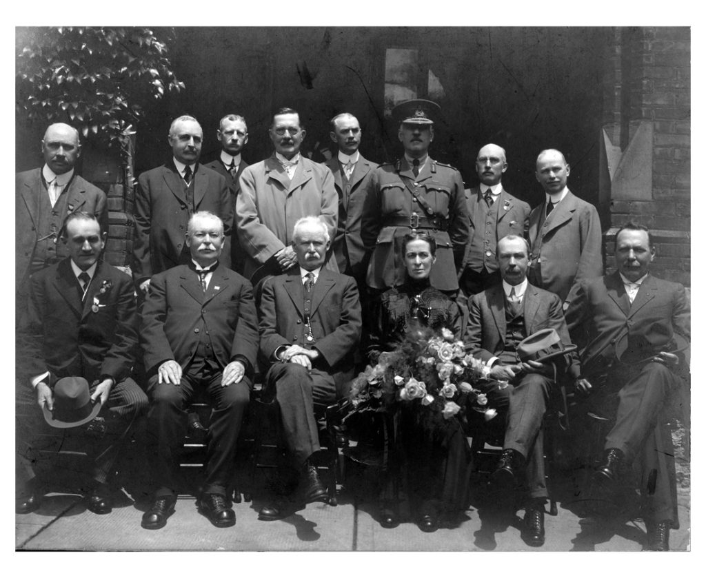 A woman with a bouquet of roses and thirteen men sit for a photo.