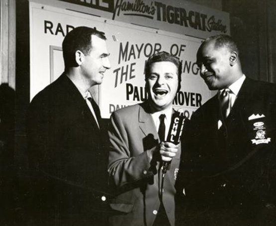 Three men gathered around a C.H.M.L radio microphone and sign Mayor of the Morning Paul Hanover.