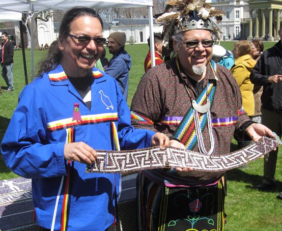 Two people wearing traditional clothing holding a belt made of wampum.