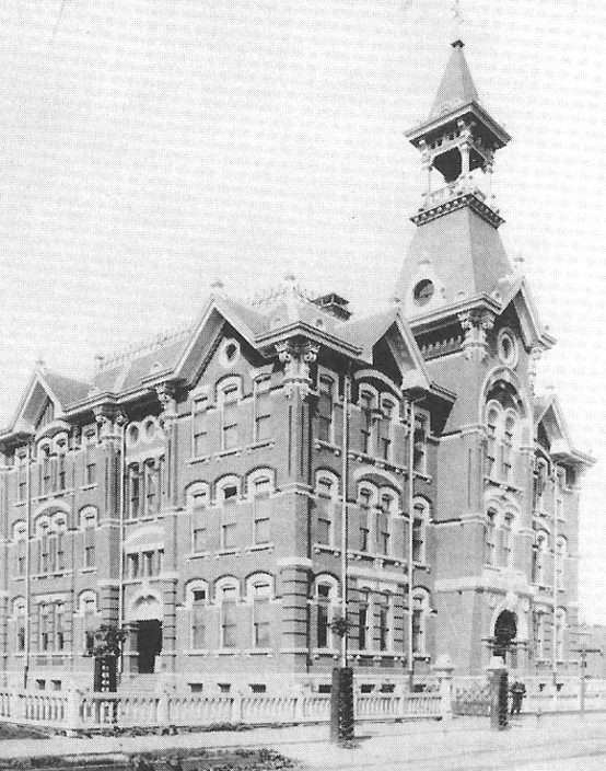 Three story boxy brick building with bell tower and spire, arched windows and ornamental roof cresting.