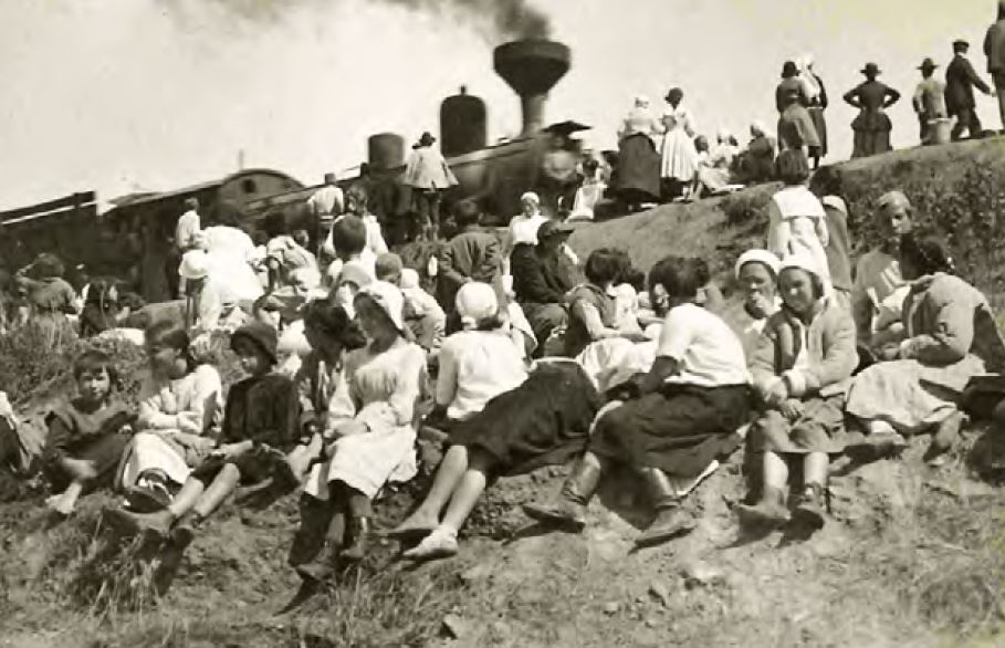 A large gathering of mostly children sitting on an embankment near a stopped steam train.