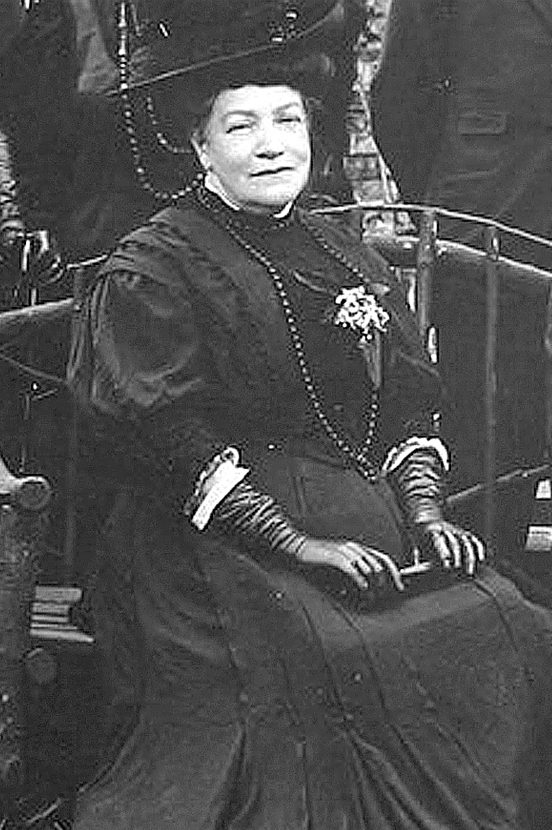 A seated woman wearing a formal long black dress, a large hat and long silk gloves characteristic of the Victorian era upper classes.