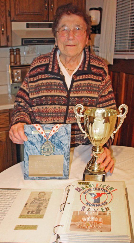 Senior woman proudly displaying a trophy, medal and scrapbook clippings on her kitchen table.