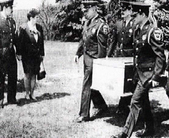 Men in uniform carry a casket while a man in uniform and woman look on.