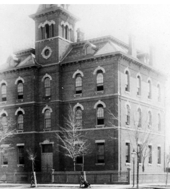 Three story boxy brick building with bell tower, arched windows and ornamental roof cresting.