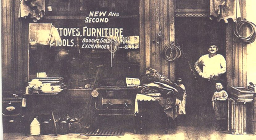 Storefront of business selling stoves, furniture and tools with adult and child in doorway.