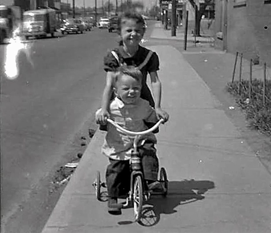 A young girl and boy ride together on a tricycle down the sidewalk of Burlington Street.