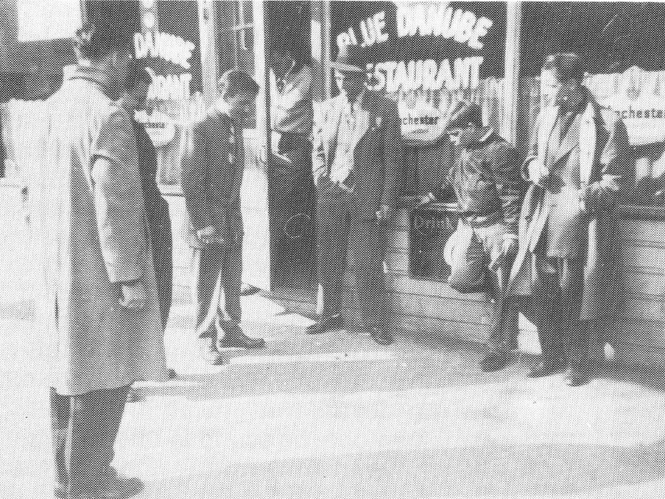 Seven men of various ages playing craps in front of the Blue Danube Restaurant.