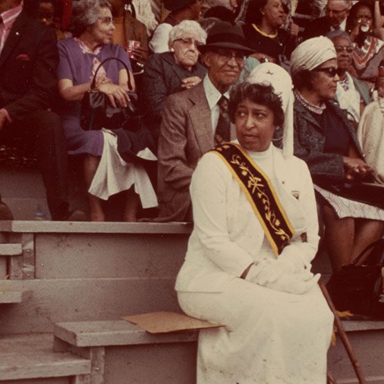 A woman dressed formally in a white dress suit with a hat and a gold and black sashe sitting in a stadium.