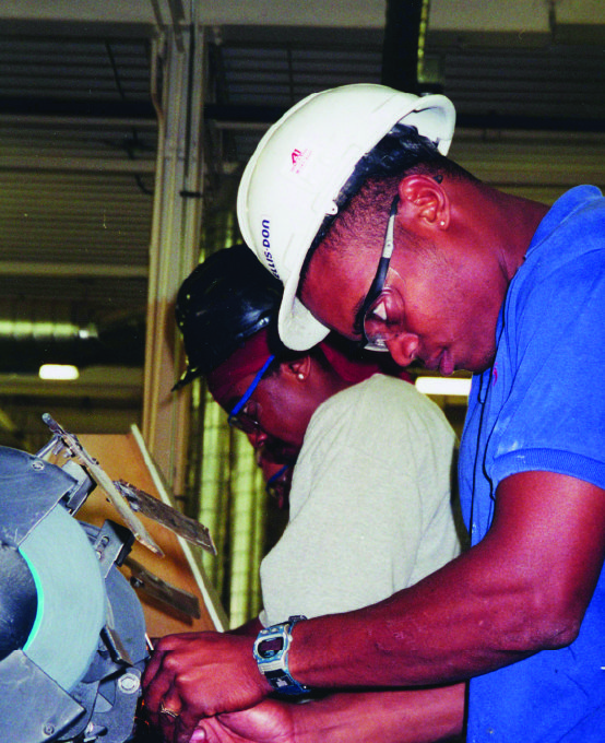 Young male workers standing at machines while wearing hard hats and safety glasses.
