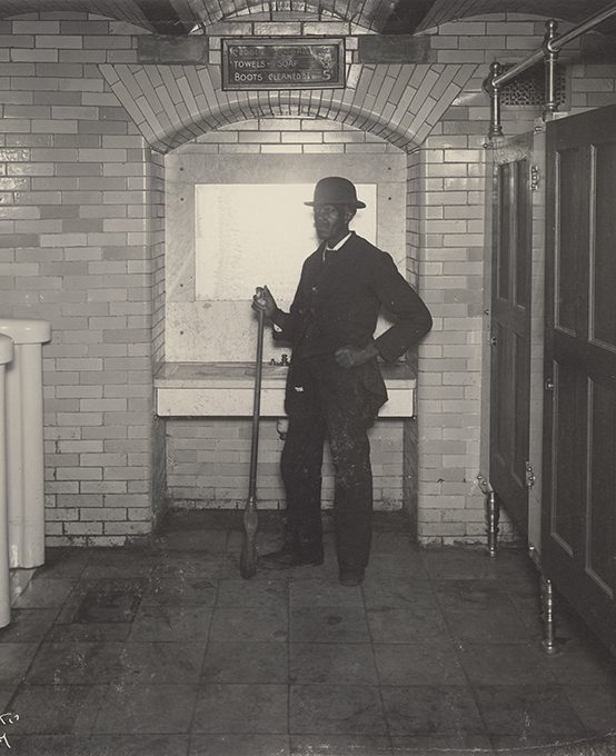 A well dressed man with a broom standing in a public washroom.
