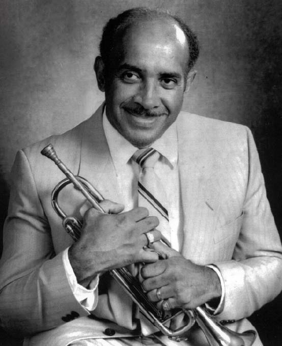 Headshot of a smiling mature man with a moustache wearing a suit and tie while holding a trumpet.
