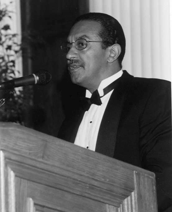 A man with moustache and glasses, in formal attire speaking into a podium microphone.