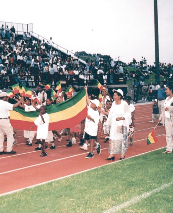 A parade of children and adults carrying a banner and mini flags with horizontal green, yellow and red stripes.