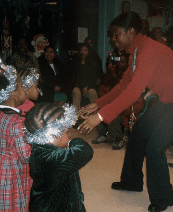 A gathering of children wearing tinsel on their heads as adults look on.