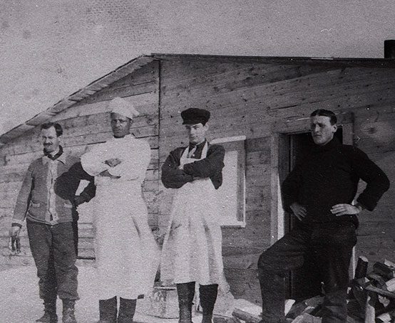 Four men standing outside a wooden cabin with one man dressed in his cook whites and hat.