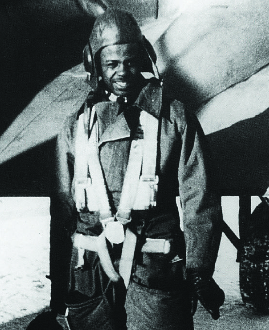 A young man wearing an aviator hat, jumpsuit and parachute harness standing beside a plane.