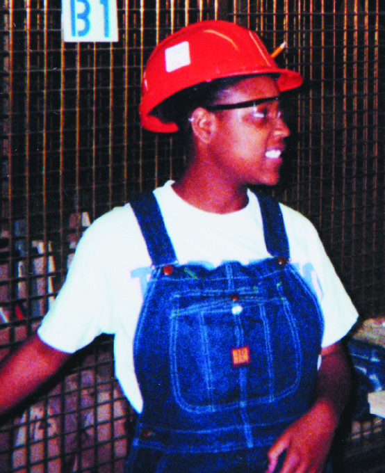 A young woman dressed in denim overalls wearing a white t-shirt and red hard hat standing in front of an metal mesh enclosure.