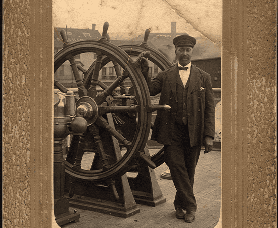 A man holding onto a ship wheel handle wearing nautical clothing and a bow tie.