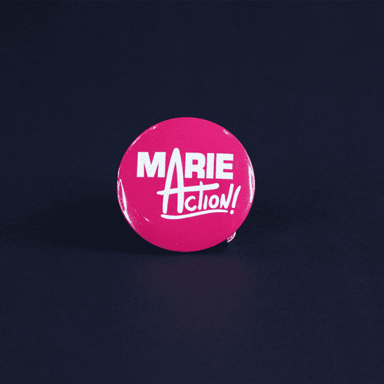 A pink lapel pin with white text showing the name Marie sharing the 'a' with the word action.