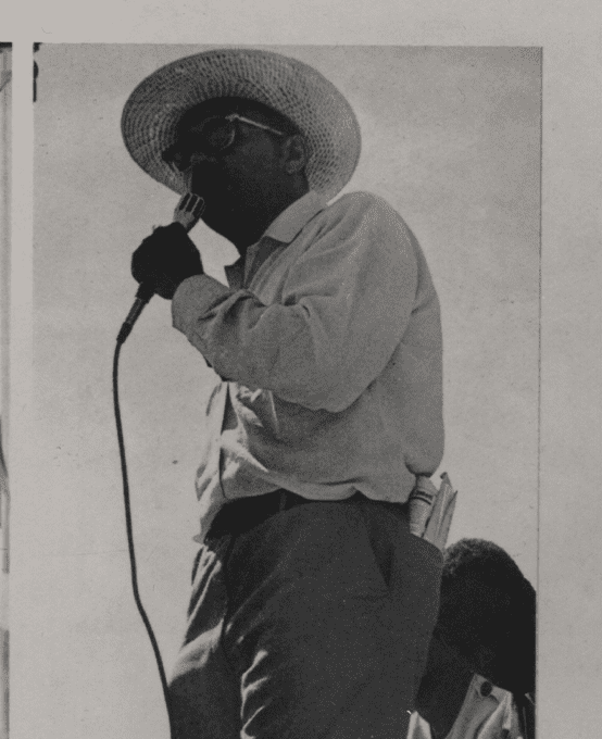 A man with glasses wearing a straw hat speaks into a microphone in his left hand.