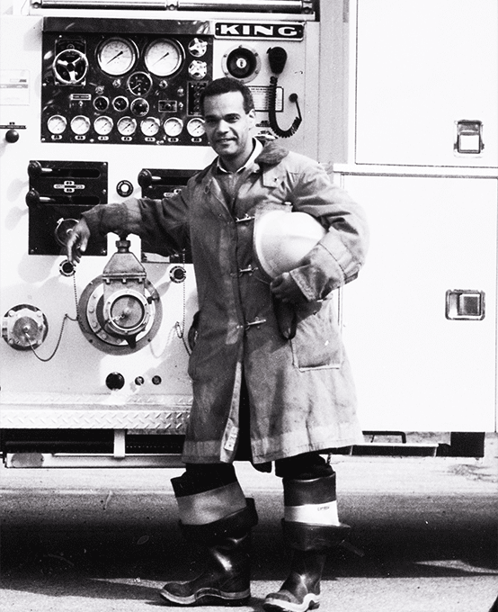 A man leans against a fire truck while dressed in his turnout gear.