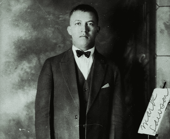 A stoic young man wearing a suit and bow tie stands in a doorway.