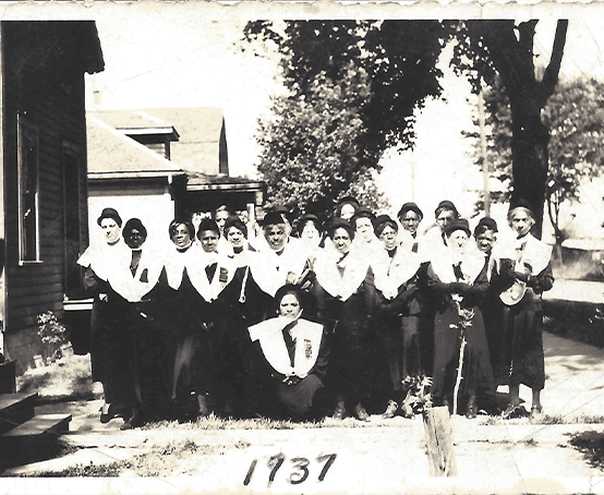 Eighteen women in black dresses with wide white shoulder coverings.