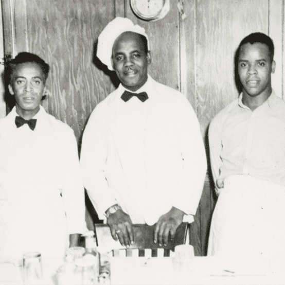 Three men dressed in white with bow ties, one is wearing a cook's hat.