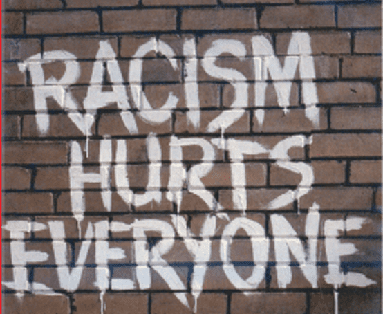 White spray paint on a red brick wall reads 'Racism Hurts Everyone'